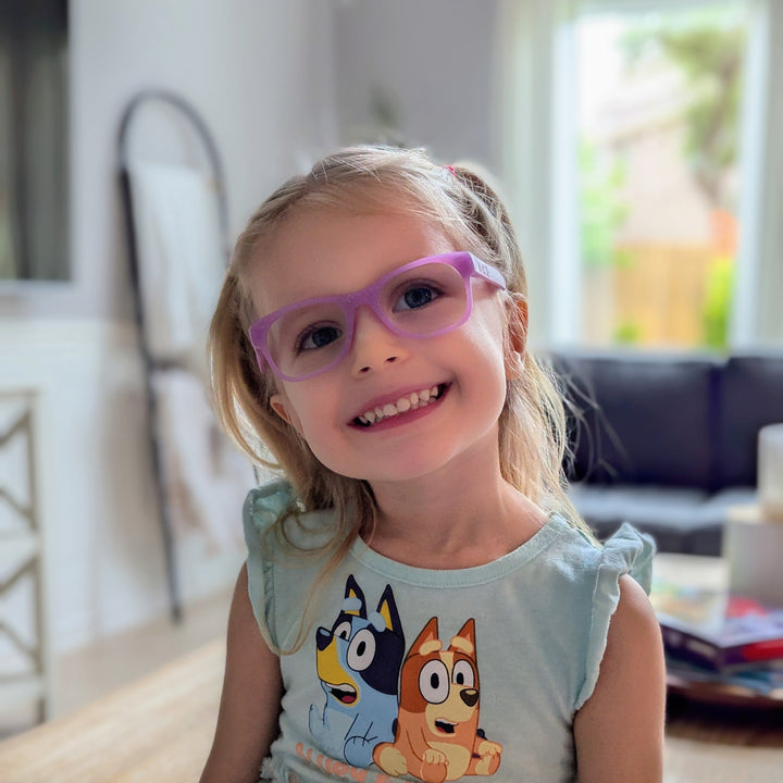 Young girl wearing purple glasses and a dress with cartoon characters indoors.