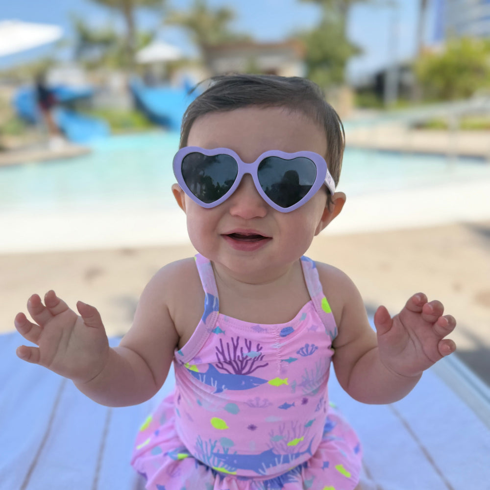 Baby in a pink swimsuit with a heart-shaped sunglasses sitting on a towel by a pool.
