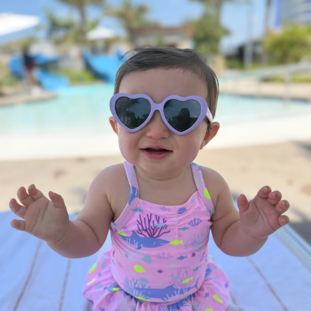 Baby in a pink swimsuit with a heart-shaped sunglasses sitting on a towel by a pool.