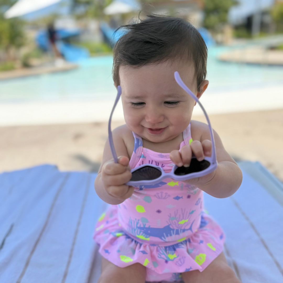 Baby in a pink swimsuit holding sunglasses at a poolside.