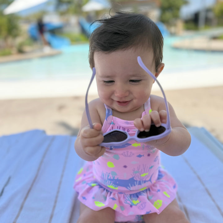 Baby in a pink swimsuit holding sunglasses at a poolside.