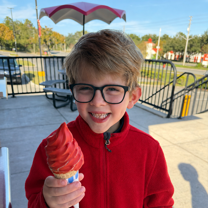 A young boy with glasses and a red fleece jacket smiles while holding a soft-serve ice cream cone outdoors on a patio.