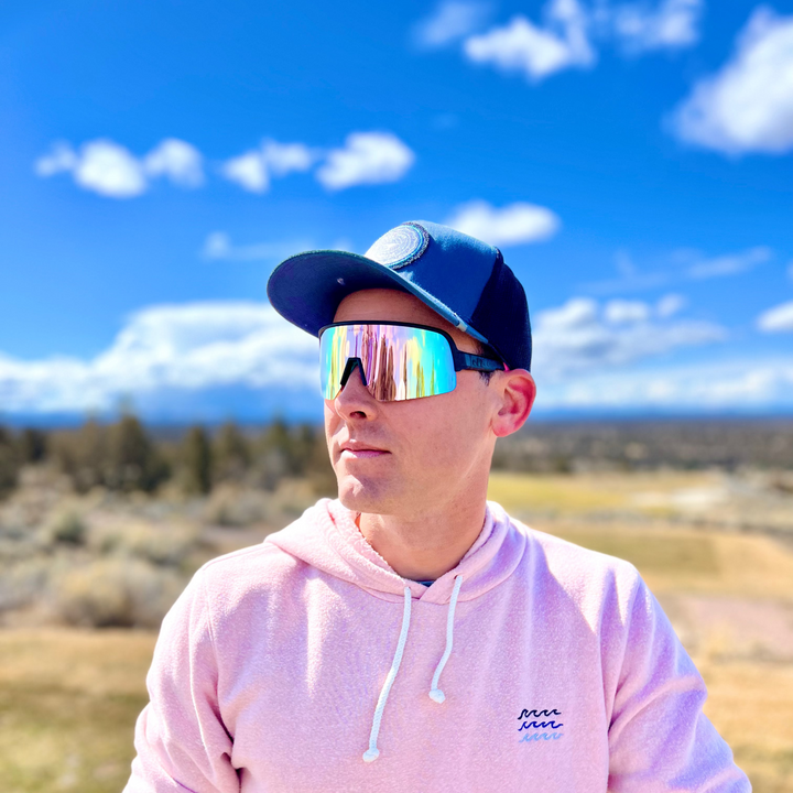 Person wearing a pink hoodie and colorful sunglasses with a blue sky and clouds in the background