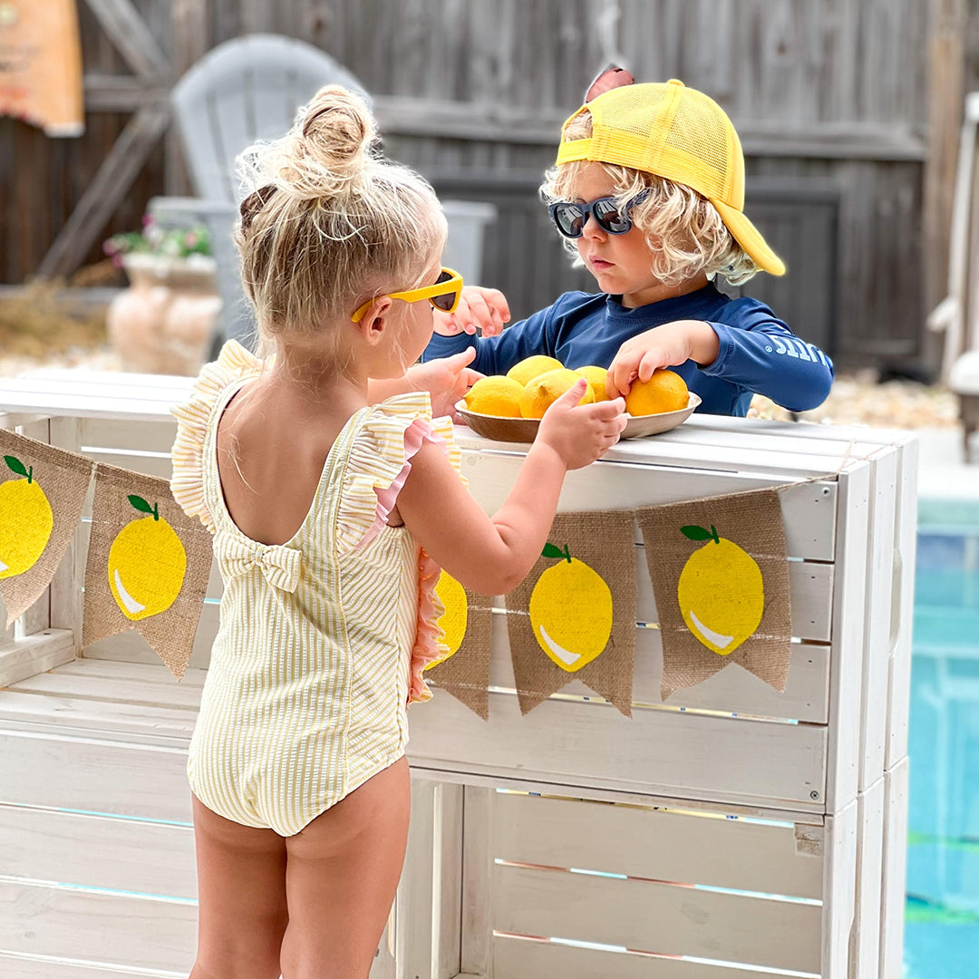 Traje de baño de sirsaca para niño pequeño de RuffleButts con gafas de sol amarillas Roshambo